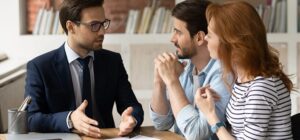 Business professional in a suit engaged in a conversation with a couple, with a focus on collaboration and discussion, seated at a table in a modern office space.