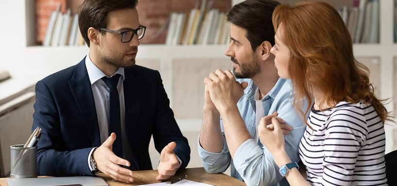Business professional in a suit engaged in a conversation with a couple, with a focus on collaboration and discussion, seated at a table in a modern office space.